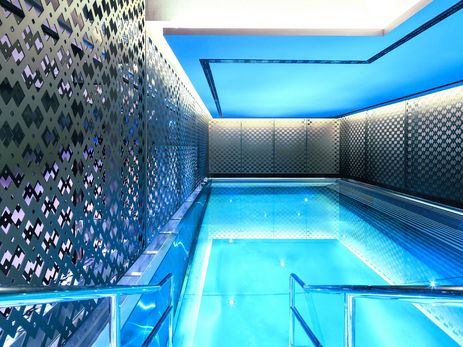 Innenansicht des luxuriösen Poolbereichs im ELEMENTS TurmCarrée in Frankfurt am Main. Ein beleuchtetes Schwimmbecken mit klarem, blauem Wasser steht im Zentrum. Die linke Wand ist mit großflächigen, dunkelgrauen Metallpaneelen verkleidet, die ein komplexes, rautenförmiges Lochmuster aufweisen und als dekorativer Sichtschutz dienen. Eine indirekte, blaue Deckenbeleuchtung verstärkt die entspannende Atmosphäre des modernen Wellnessbereichs. Treppengeländer führen sicher in das Wasser.
