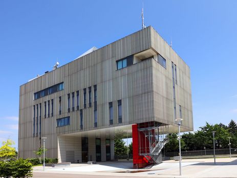 Eine Außenansicht der Integrierten Leitstelle in Karlsruhe von der Seite. Das Gebäude präsentiert sich als markanter, hellgrauer Block aus Aluminium-Paneelen, der über einem zurückgesetzten Erdgeschoss aus Sichtbeton zu schweben scheint. An der Längsseite sind schmale, vertikale Lichtschlitze sowie ein horizontales Fensterband bündig in die Metallhaut integriert. Die leuchtend rote Stahltreppe unter dem Auskragungsbereich setzt einen starken farblichen Akzent in der ansonsten reduzierten Materialpalette.